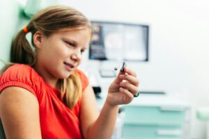 A young girl looks at her hearing aid with a look of fascination on her face. She's white with dark blonde hair which is tied back in pigtails and she's wearing a bright red tshirt.