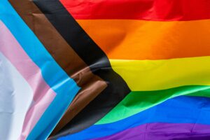 The Progress Pride Flag. It shows the traditional pride stripes, with white, pink, and light blue stripe to represent the Trans community, and black and brown stripes representing communities of colour, the black strip in particular representing the thousands of individuals that the community list during the HIV/AIDS crisis in the 1980s and 1990s.