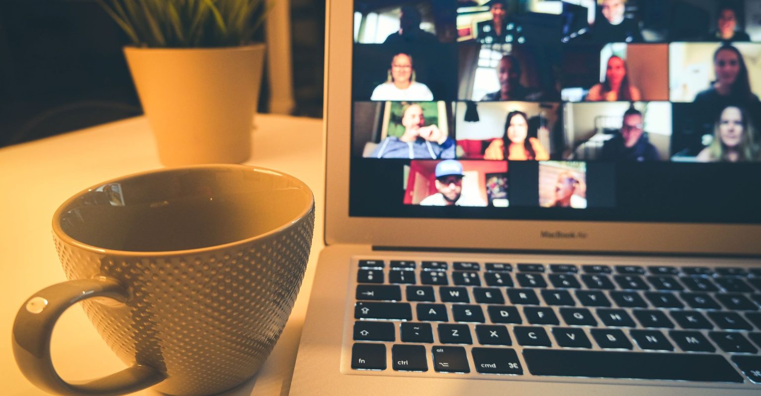 A laptop sitting on a table next to a mug of tea. The laptop screen is showing a Zoom meeting with multiple participants.