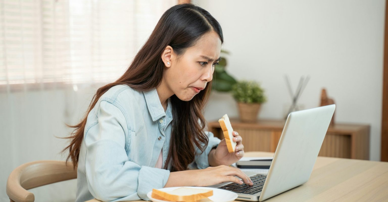 A woman is working at a laptop while eating breakfast. She looks a bit harassed.