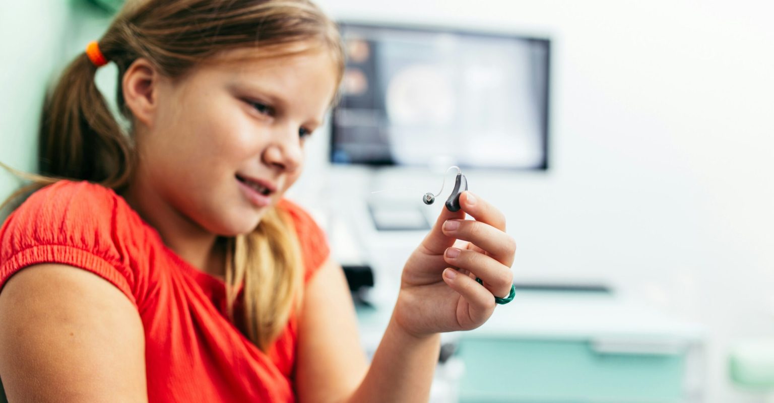 A young girl looks at her hearing aid with a look of fascination on her face. She's white with dark blonde hair which is tied back in pigtails and she's wearing a bright red tshirt.